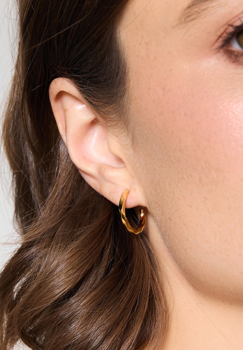 Złota skręcona kolczyka na uchu, z gładką fakturą i warstwowym wzornictwem, odbijająca światło dzięki połyskliwej powierzchni.