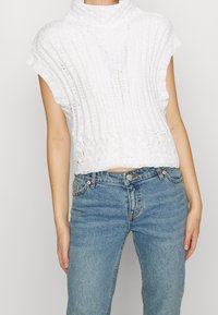 Bílý pletený crop top s vysokým límcem, s žebrovanou texturou a krátkými rukávy, spárovaný se světle modrými džíny s vysokým pasem.