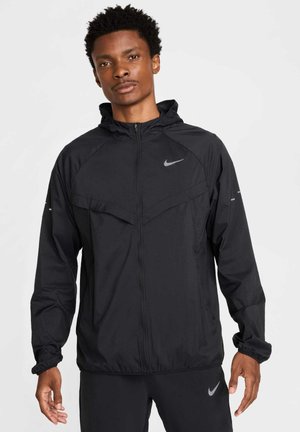 Fekete Nike széldzseki, cipzárral elöl, kapucnival és elasztikus mandzsettákkal. Két mellzsebbel és egy fényvisszaverő logóval rendelkezik a mellkasán.