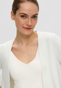 Witte gebreide cardigan met een open voorkant, gedragen over een witte V-hals top. Gladde textuur met een lichte glans en korte mouwen.