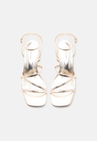 Ζευγάρι λαμπερών μεταλλικών χρυσών σανδαλιών με λουράκια, τετράγωνες μύτες και λεπτά λουράκια στον αστράγαλο σε λευκό φόντο.