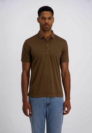 Man die een bruin poloshirt met korte mouwen en lichtblauwe spijkerbroek draagt, staand tegen een effen witte achtergrond en vooruitkijkend.