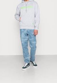 Felpa grigia con logo verde, tasca frontale e cordini; abbinata a jeans azzurri chiari e sneakers verde/bianco con design alto.