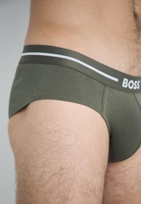 Green cotton briefs with a contrasting white and light blue striped waistband, featuring a logo at the front. Smooth texture.