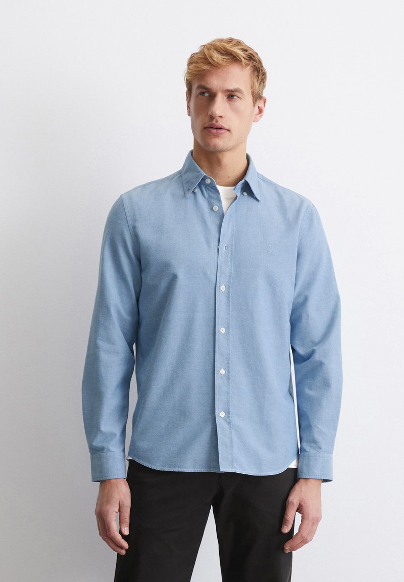Light blue long-sleeve button-up shirt with a collar, white buttons, and a subtle embroidered detail on the chest. Worn over a white shirt.