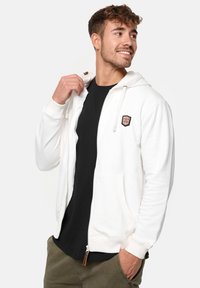 Weißer Zip-Hoodie mit einem strukturierten Logo-Patch und Kordelzugkapuze; getragen über einem schwarzen T-Shirt und kombiniert mit khakifarbenen Shorts.