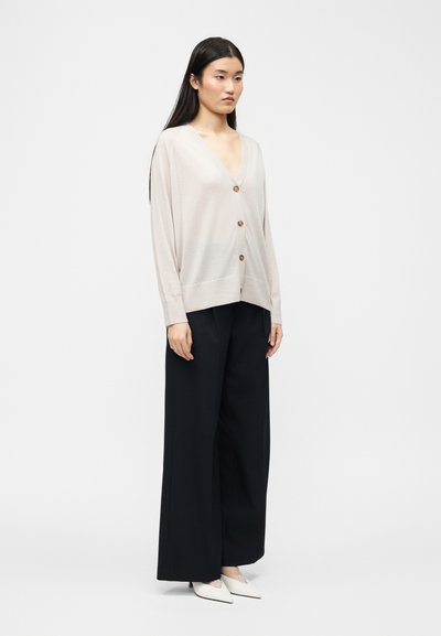 Γυναίκα που στέκεται, φοράει ανοιχτό μπεζ ζακέτα με κουμπιά, μαύρο παντελόνι φαρδύ και λευκά παπούτσια με μυτερή μύτη, μπροστά από απλό φόντο.