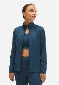 Chaqueta deportiva de color verde azulado con cremallera y cuello alto, de tejido ligero, estilo ajustado y detalles en las costuras. Llevada sobre un sujetador deportivo a juego.