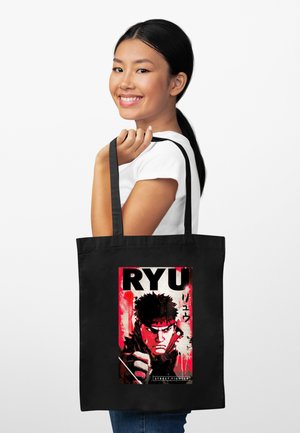 Jonge vrouw die lacht en een zwarte tas vasthoudt met een rood-zwarte afbeelding van Street Fighter-personage Ryu en Japanse tekst.