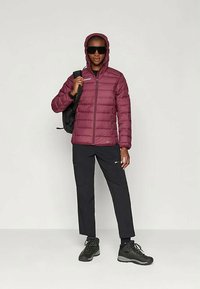 Μαρόν φουσκωτό μπουφάν με κουκούλα, κλείσιμο με φερμουάρ και ραφές τύπου καπιτονέ· συνδυασμένο με μαύρα παντελόνια και μποτάκια για πεζοπορία. Συμπεριλαμβάνονται σακίδιο πλάτης και γυαλιά ηλίου.