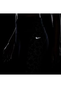 Czarne legginsy sportowe z teksturowanym wzorem i odblaskowym logo Nike po boku. Zbliżenie z ciemnym tłem.