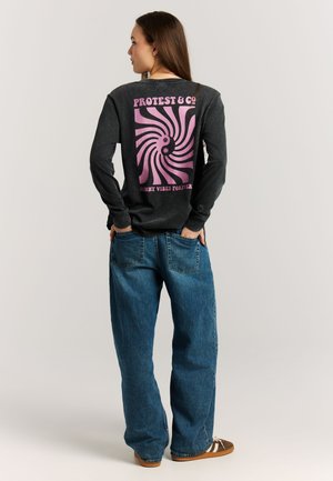 Vrouw draagt een zwart shirt met lange mouwen met een roze wervelend ontwerp en tekst, gecombineerd met losse blauwe spijkerbroeken en bruine sneakers, staand met handen in de zakken.