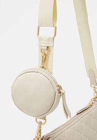 Beige Tasche mit gestepptem Muster, ausgestattet mit einem abnehmbaren, runden Täschchen. Goldfarbene Hardware-Akzente und ein breiter Stoffriemen sind sichtbar.