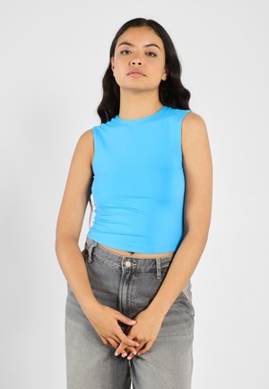 Top corto sin mangas azul en una tela suave y elástica, combinado con unos vaqueros de mezclilla gris de tiro alto, que cuentan con un cierre de botón y cremallera.