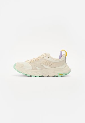 Bežs sporta apavas ar strukturētu acs materiālu un zamša pārklājumiem, zaļa gumijas zole, lilā aizmugures akcents un dzeltena cilpa. Iespējams, ar Vibram zīmolu.