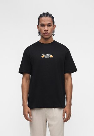 Νέος άνδρας με πλεξίδες φοράει ένα μαύρο T-shirt με το κείμενο "Valencia Naranjo Verde" και πορτοκαλί γραφικά, συνδυασμένο με ανοιχτό μπεζ παντελόνι.