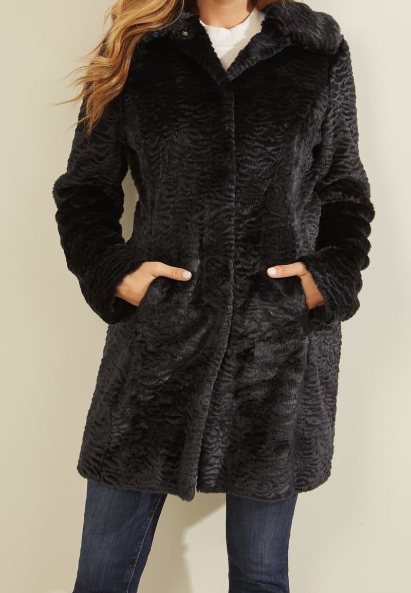 Abrigo negro de piel sintética con un patrón ondulado texturizado, cuello abotonado, bolsillos laterales y mangas largas, llevado sobre unos vaqueros.