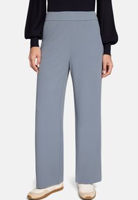 Pantalon gris clair à jambes larges réalisé en tissu lisse. Il présente une taille haute et une silhouette nette et ajustée, porté avec des baskets blanches.