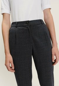 Person trägt dunkelgraue karierte Hosen mit einem schwarzen Knopf und Falten, kombiniert mit einem weißen Kurzarm-Oberteil, die Hände an den Seiten ruhend.