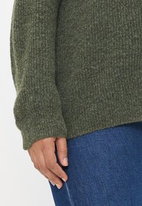 Suéter de punto acanalado verde con mangas largas, textura suave y corte relajado, combinado con jeans azul oscuro. Incluye un puño visible.