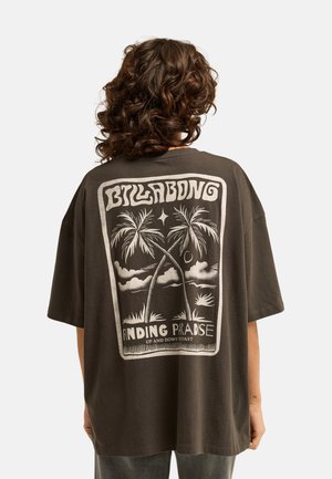 Osoba o kręconych włosach nosząca ciemny, oversize'owy t-shirt z grafiką palmy i tekstem "Billabong Finding Paradise" na plecach.