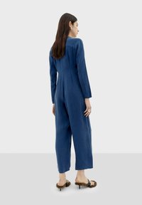 Navyblauwe jumpsuit met lange mouwen en rechte snit, met een detail van achternaad. Combineert met zwarte hakken en pluizige accenten.