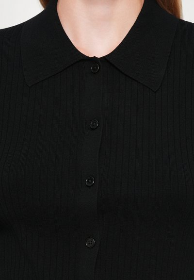 Musta ribattum shirt, jossa on kauluskanta ja viisi mustaa nappia. Materiaali on sileä ja tiukka, korostaen hoikkaa siluettia.