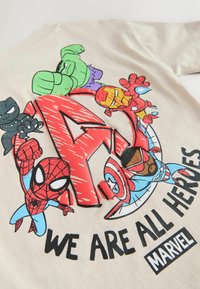 Bež majica z barvitim stripovskim motivom superjunakov okoli velike rdeče črke 'A' z napisom »MI SMO VSI JUNAKI« in logotipom Marvel.