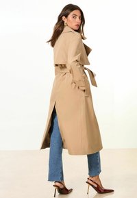 Trench beige con cintura ceñida, aberturas laterales y cuello alto. Combinado con jeans de dobladillo deshilachado y tacones de charol.