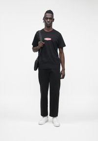 Melnā T-krekls ar sarkanu "Guess Jeans" logotipu, kopā ar melnām biksēm un baltām sporta kurpēm. Modelis nēsā melnu plecu somu, stāv taisni.