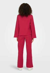 Completo pantalone a campana in tessuto rosa profondo e testurizzato. Maniche lunghe e top a vestibilità ampia. Linee pulite con un design semplice. Sneakers bianche visibili.