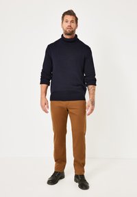 Marinefarbener Rollkragenpullover mit gerippten Bündchen und Saum, kombiniert mit beige Hosen und schwarzen Schuhen. Modell steht vor einem weißen Hintergrund.