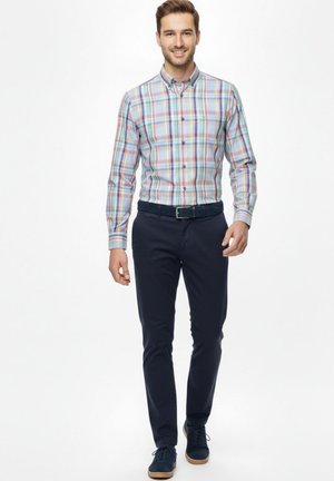 Hombre caminando hacia adelante vistiendo una camisa abotonada de cuadros multicolores, pantalones azul marino oscuro, cinturón negro y zapatos oscuros con suelas claras.