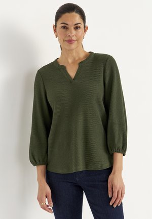 Vrouw met donker haar naar achteren gebonden, die kleine gouden oorbellen draagt en een olijfgroene, getextureerde top met lange mouwen en donkere spijkerbroek.