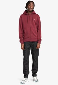 Felpa con zip color marrone bordeaux, dotata di cappuccio con cordino, tasche frontali e polsini a coste, abbinata a jeans di denim scuro e sneakers argentate.