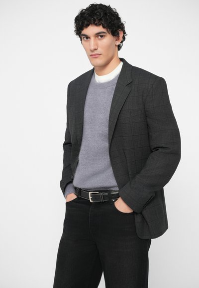 Junger Mann mit lockigem Haar, der ein dunkelkarriertes Sakko, einen grauen Pullover über einem wei�ßen Hemd, schwarze Jeans trägt und die Hände in den Taschen hat.