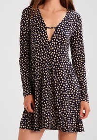 Kvinna som bär en marinblå långärmad klänning med beige cirkulära mönster och en djup V-ringning med ett litet horisontellt band.