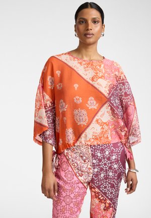 Vrouw draagt een losse, patroonrijke oranje, roze en paarse top en bijpassende broek, staand tegen een effen lichte achtergrond.