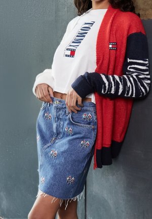 Kobieta ubrana w biały krótki top Tommy Jeans, czerwono-granatowy kardigan oraz dżinsową spódnicę z haftowanymi wzorami flag.