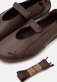 Chaussures Puma en cuir marron style Mary Jane avec des brides Velcro et une paire de lacets plats marron assortis sur fond blanc.