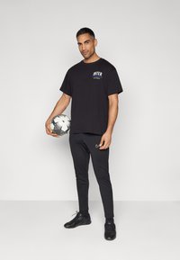 Svart t-skjorte med "INTER"-logo på brystet, kombinert med svarte treningsbukser og svarte sko. En fotball holdes i den ene hånden.