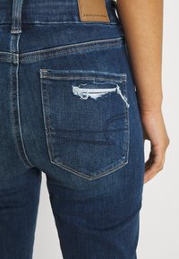 Mörkblå denimjeans med sliten bakficka, med en läderlogga vid midjan och orangea sömnadsdetaljer.