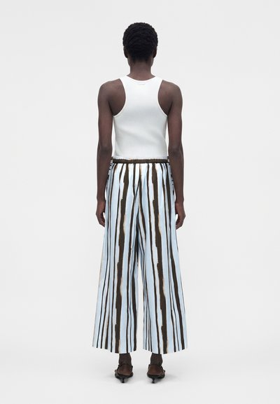 Άτομο με κοντά σγουρά μαλλιά που φοράει λευκό αμάνικο τοπ, παντελόνι με φαρδιά μπατζάκια και κάθετες μαύρες και λευκές ρίγες, και μαύρα σανδάλια με τακούνι, με την πλάτη προς τον θεατή.