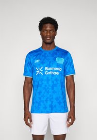 Man wearing blue Bayer Leverkusen football jersey with sponsor logos and white shorts, standing against plain background.