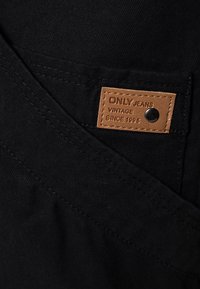 Jeans neri con un tessuto testurizzato, dotati di un'etichetta in pelle marrone chiaro impressa con "ONLY JEANS VINTAGE SINCE 1995" e un rivetto metallico.