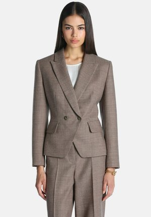 Kvinde med glat mørkt hår iført en brun skræddersyet dobbeltradet blazer og matchende bukser, stående mod en ensfarvet baggrund.