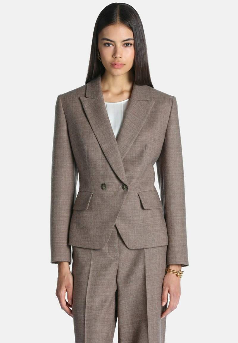 Femme aux cheveux longs et raides de couleur foncée, portant un blazer marron croisé taillé sur mesure et un pantalon assorti, se tenant devant un fond uni.