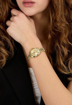 Frau mit lockigem Haar und goldener Armbanduhr, die ihre Hand am Kinn abstützt, gekleidet in einem schwarzen Blazer und weißem Top.