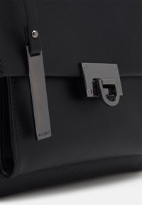 Svart texturerad läderhandväska med ett stort metalliskt spänne med ett cirkulärt handtag, embossed logotagg och släta detaljer.