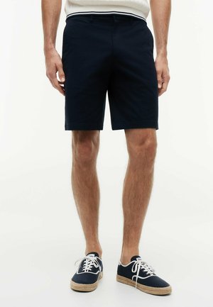 Mand iført marineblå shorts, marineblå lærreds snøresko med beige såler og en hvid top, stående på en ensartet hvid baggrund.